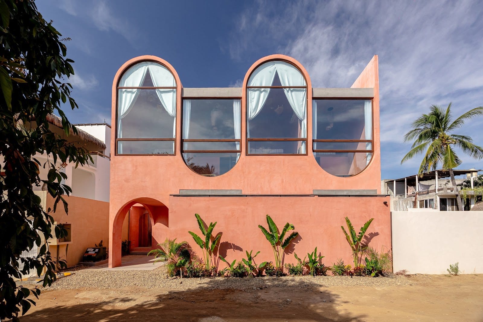 Street-facing pink facade of Amplitude Coliving with tall arched windows, tropical plants, and a palm tree.