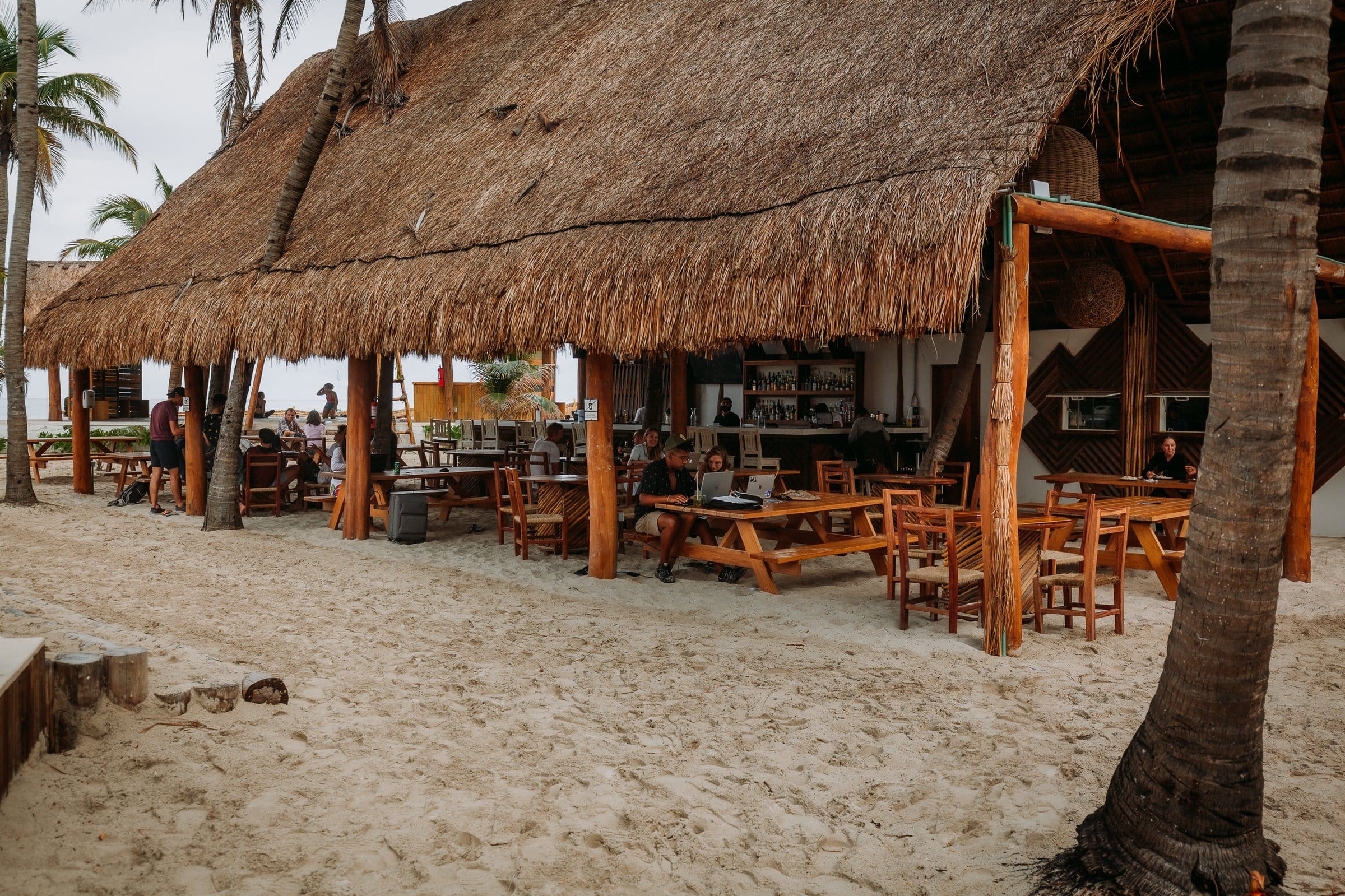 Guests socializing and working at the beachfront palapa bar and restaurant at Selina Isla Mujeres with the Caribbean sea visible in the background