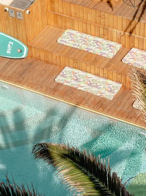 Wood deck steps with colorful cushions near clear pool water and edges of tropical plants visible.