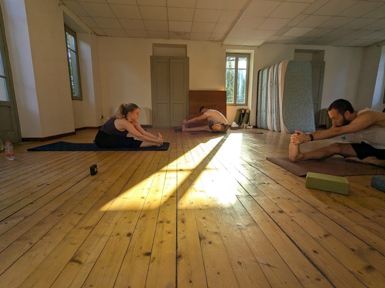 Indoor large room with wooden floors and white walls, three people stretching on yoga mats with mattresses stacked to the side.