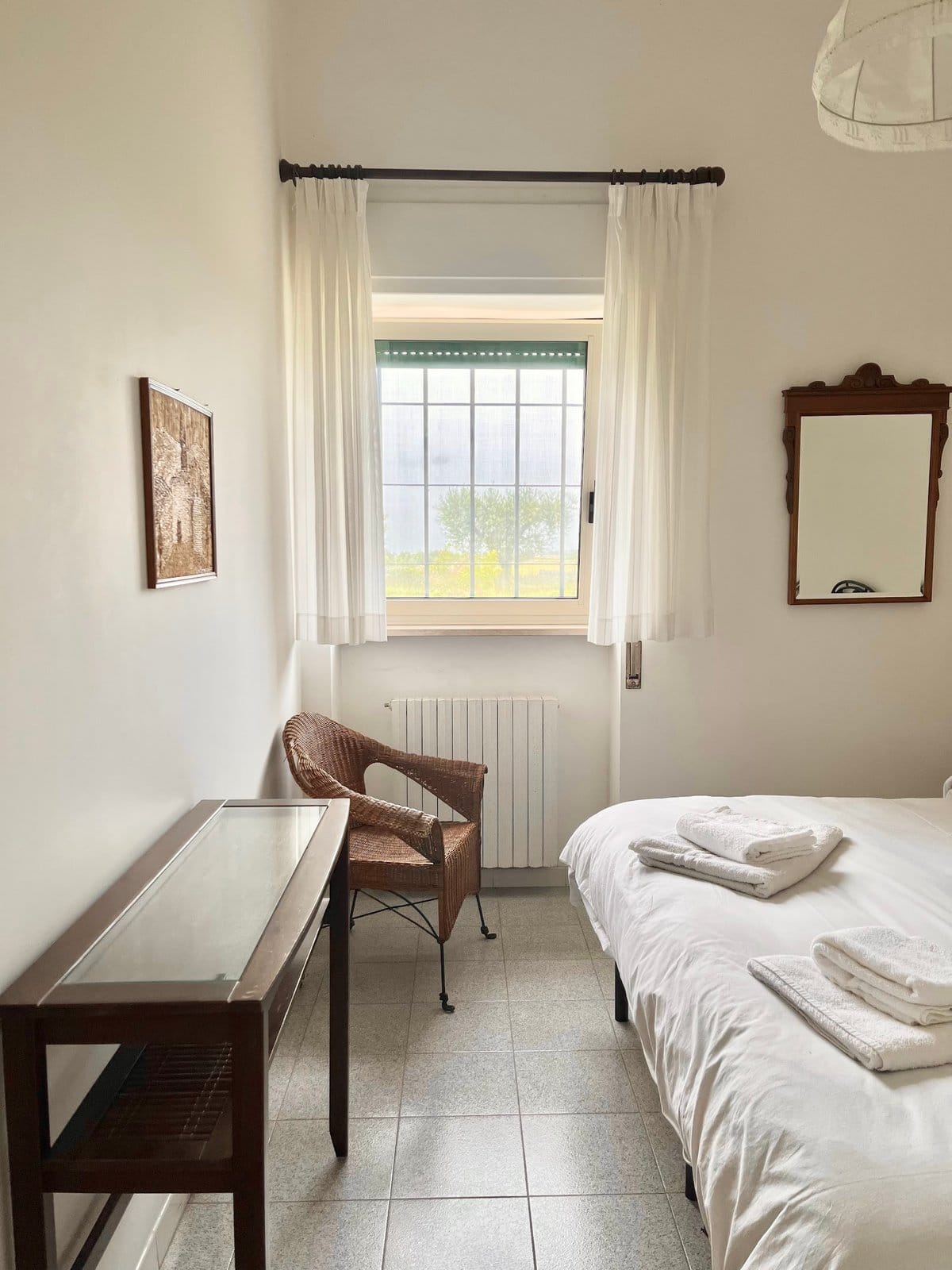 Simple bedroom with unmade bed, folded towels, wicker chair, window with curtains allowing natural light.