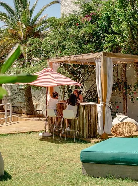 Outdoor garden seating with three people chatting under red striped umbrella in shade.