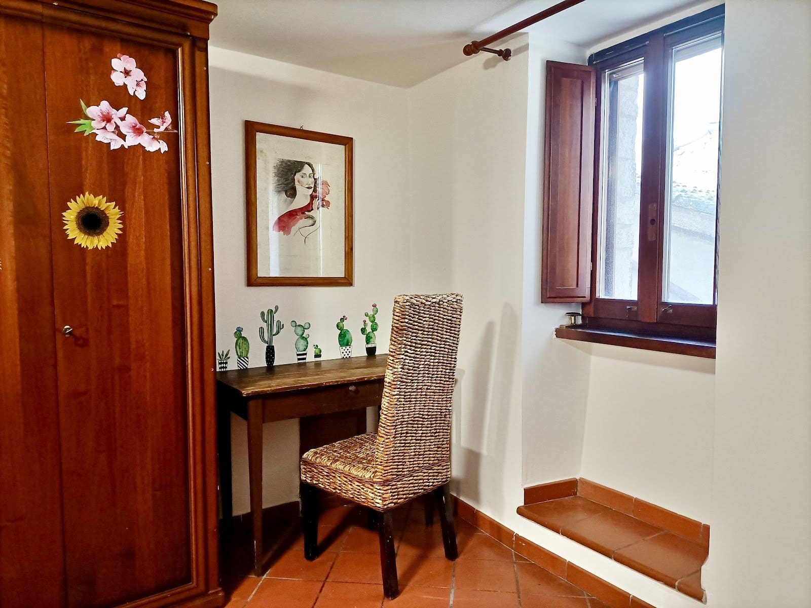A small desk with a wicker chair against a white wall decorated with cactus figurines and a drawing, wooden furniture and a window.
