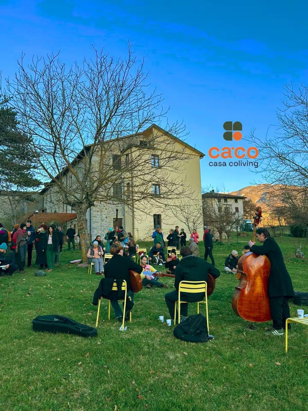 People socializing outdoors on lawn with several guitars and a double bass player performing, in front of a stone building with leafless trees.