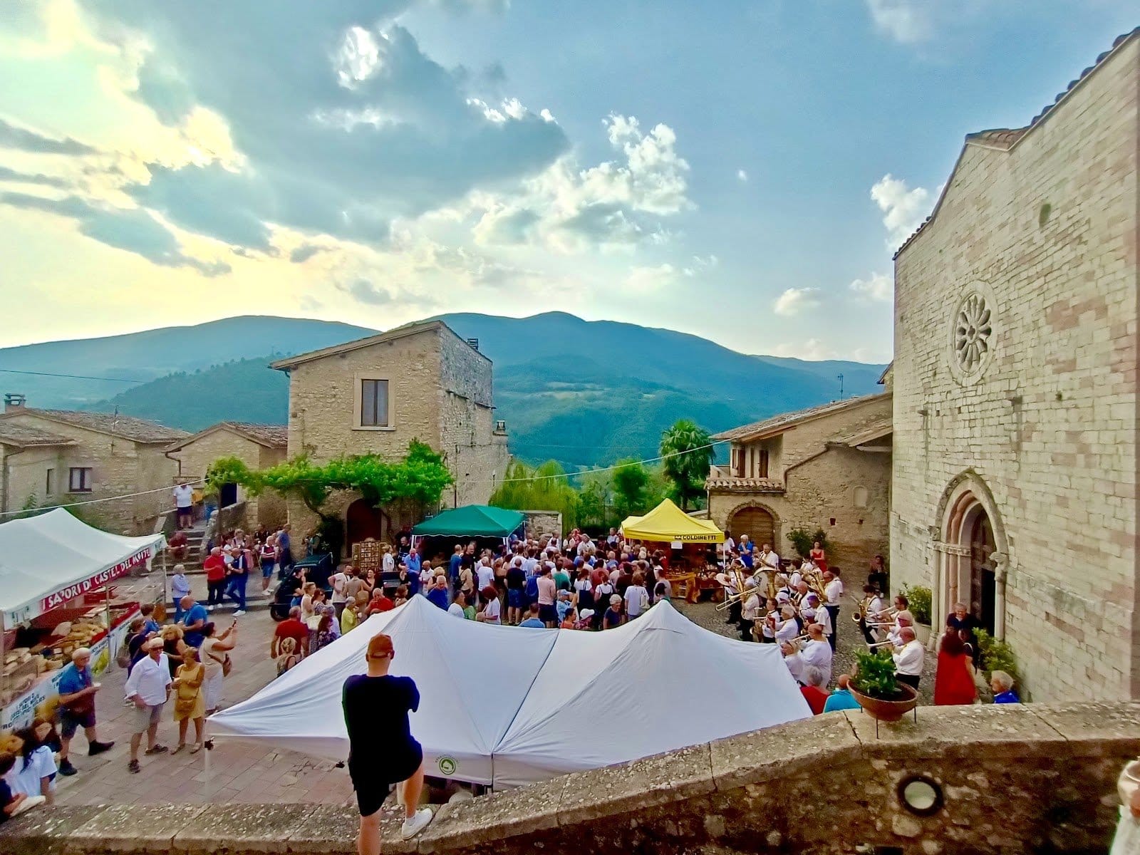 Large outdoor crowd gathered for an event in a stone paved plaza with historic stone buildings.
