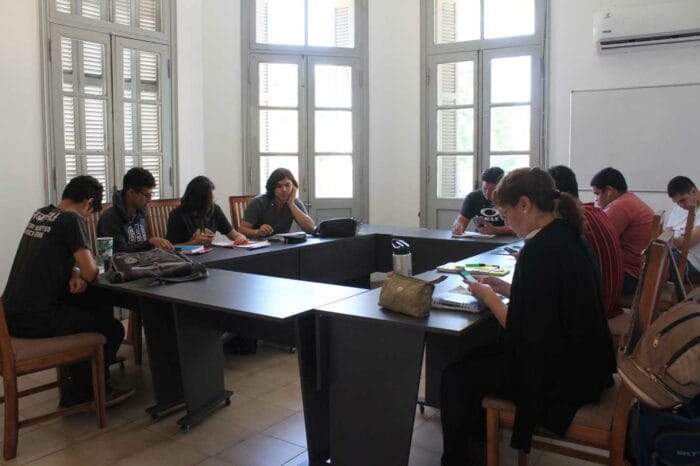 People are seated at desks arranged in a U-shape with laptops and papers, inside a bright room with tall windows.