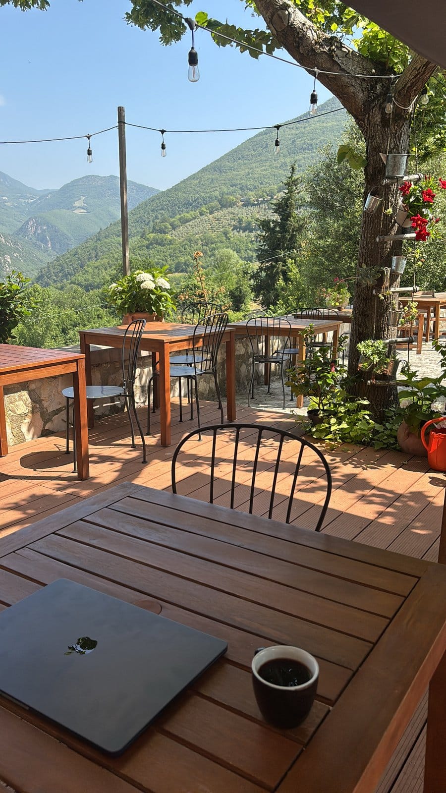 Outdoor terrace with wooden tables and chairs decorated with hanging lights, overlooking green hills and mountains.