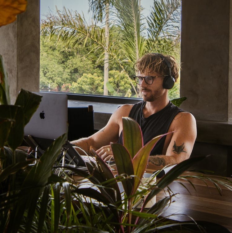 Man working on a laptop at an indoor desk surrounded by plants, with headphones and glasses, window behind him showing trees.