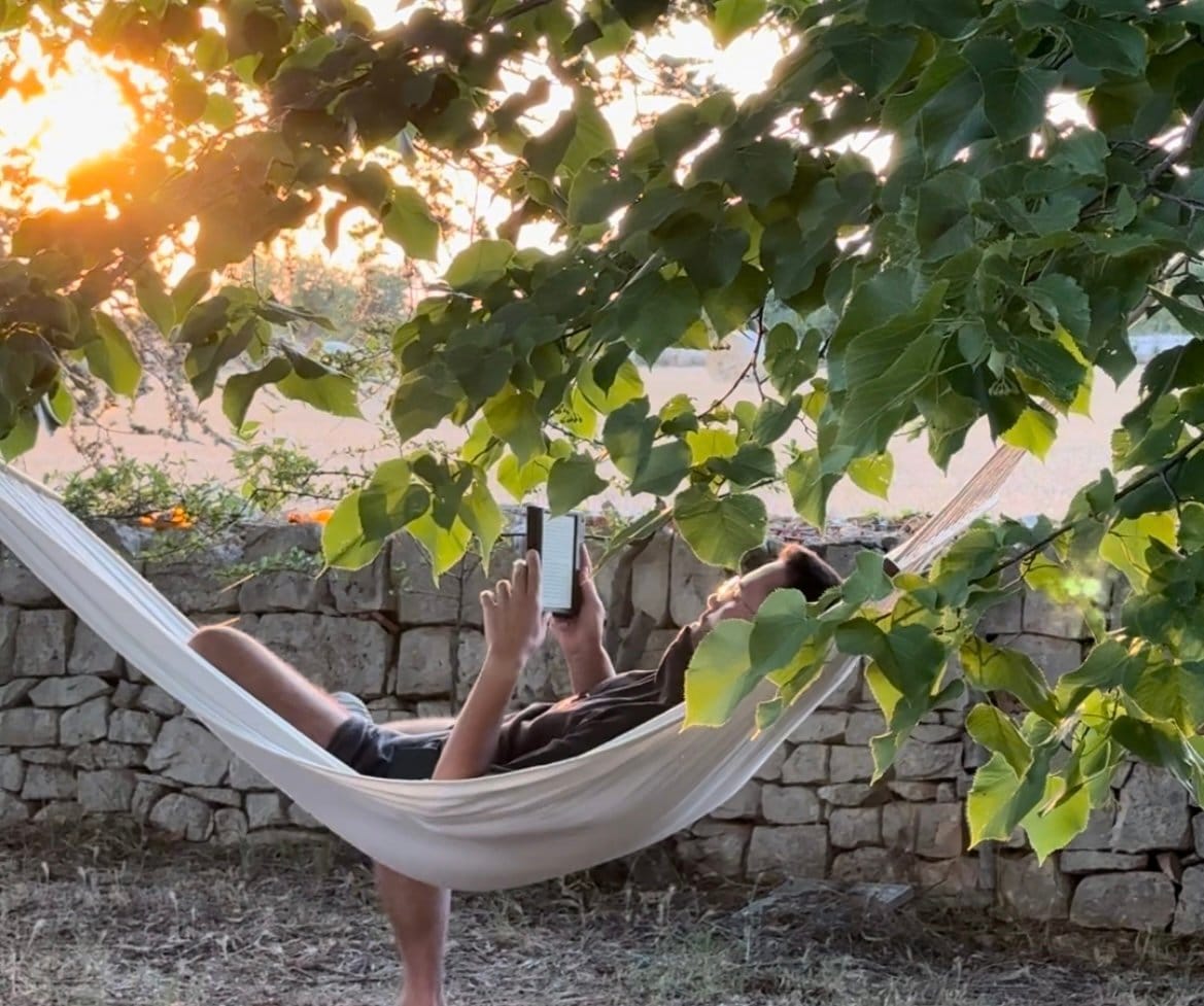 Person reclining in white hammock outdoors surrounded by trees during golden hour, reading a device.