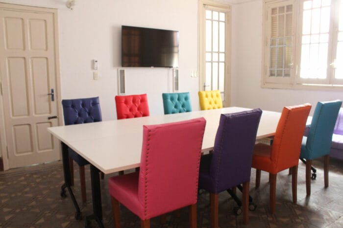 Bright room with white rectangular table and colorful chairs, TV mounted on wall visible.