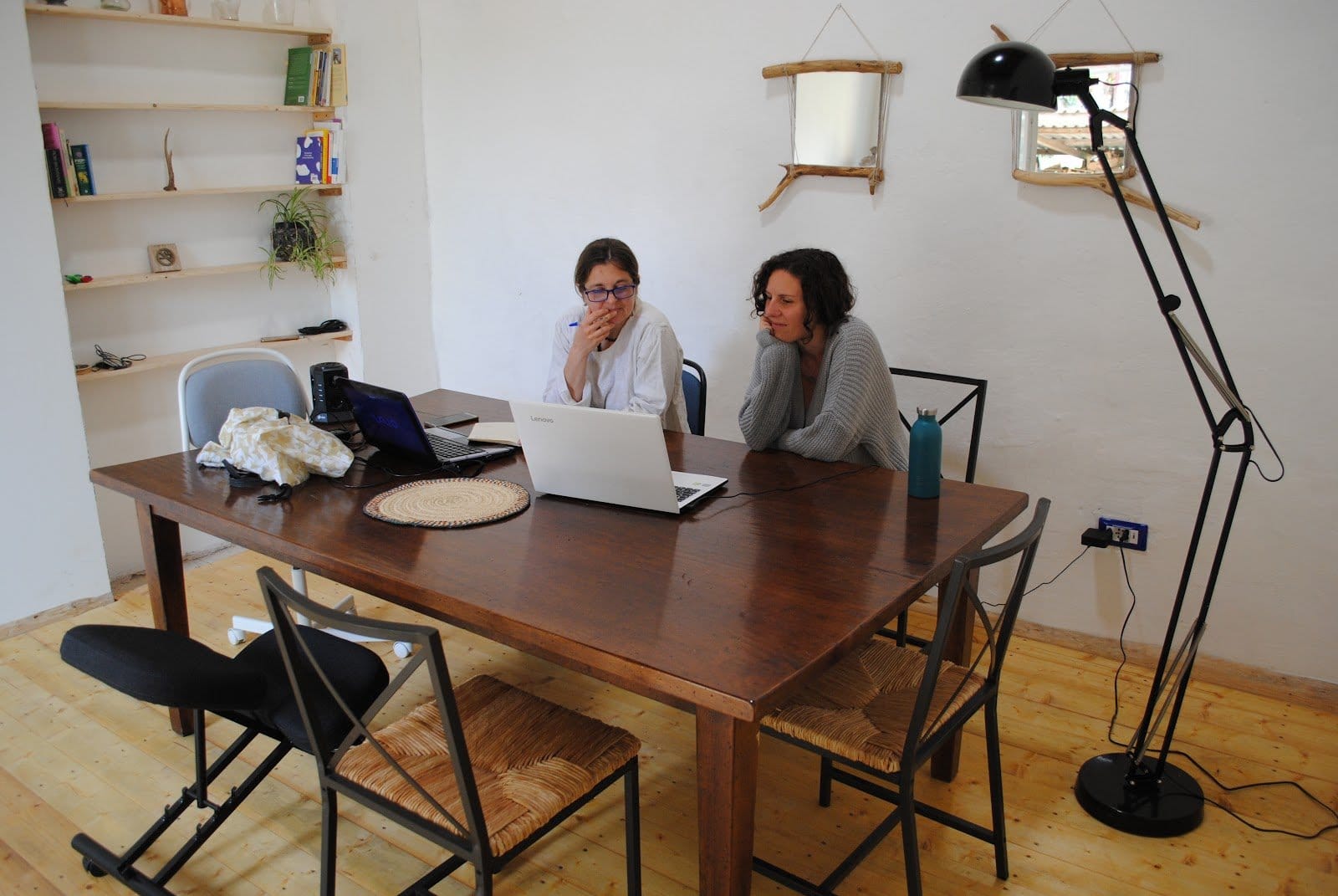 Two women sitting inside at a large wooden table with laptops, focused on work in a bright room.
