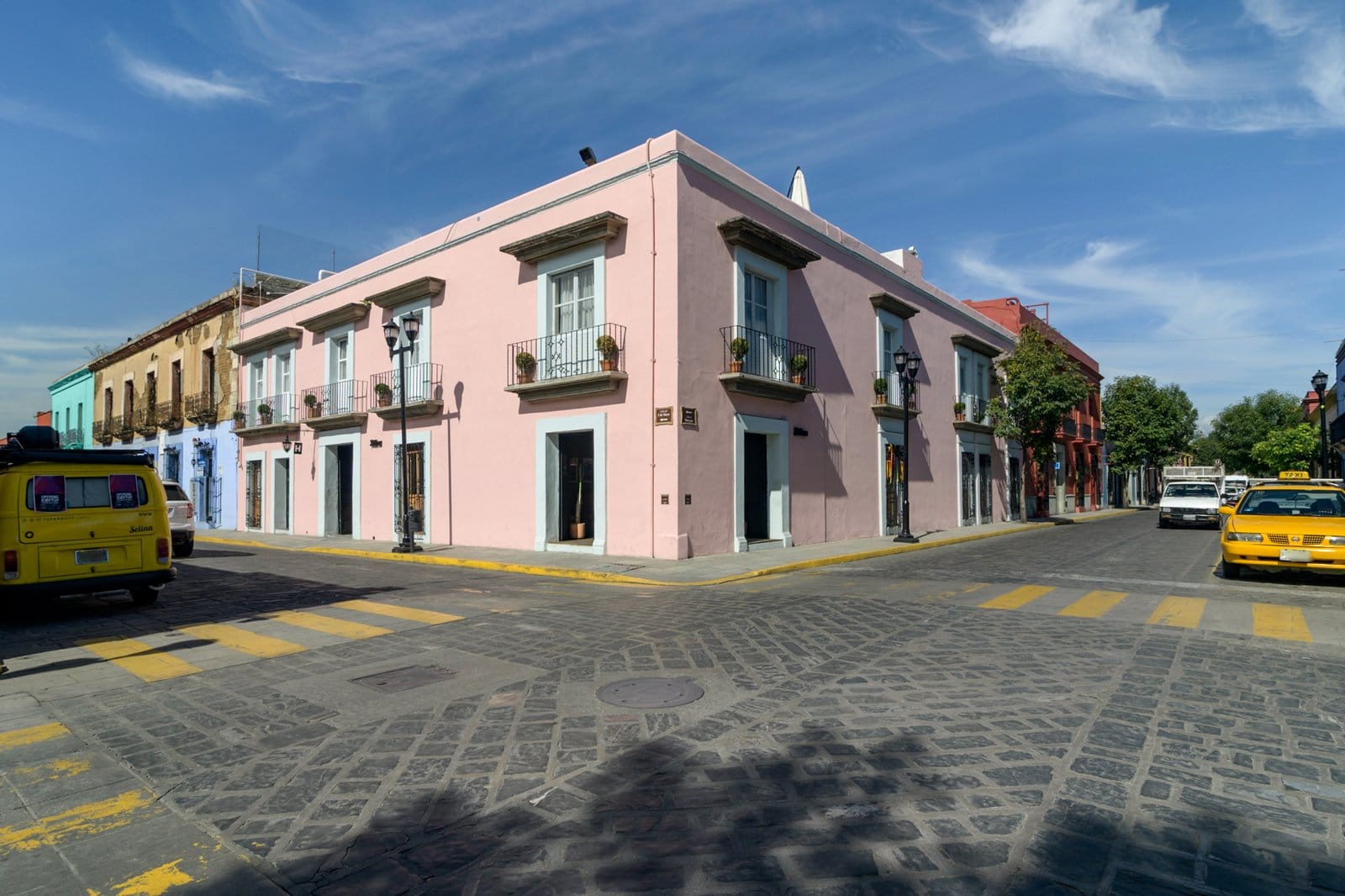 The pink colonial corner building of Selina Oaxaca on a cobblestone intersection with colorful neighboring buildings and yellow taxi