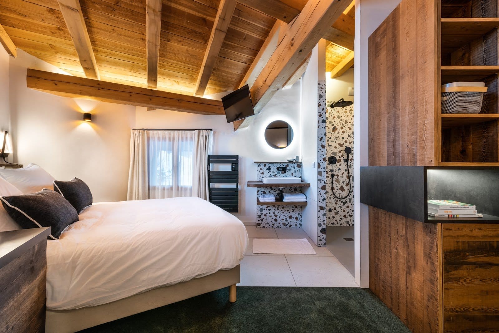 Well-lit bedroom with a white bed, green carpet, wooden ceiling beams, and a visible modern bathroom area.