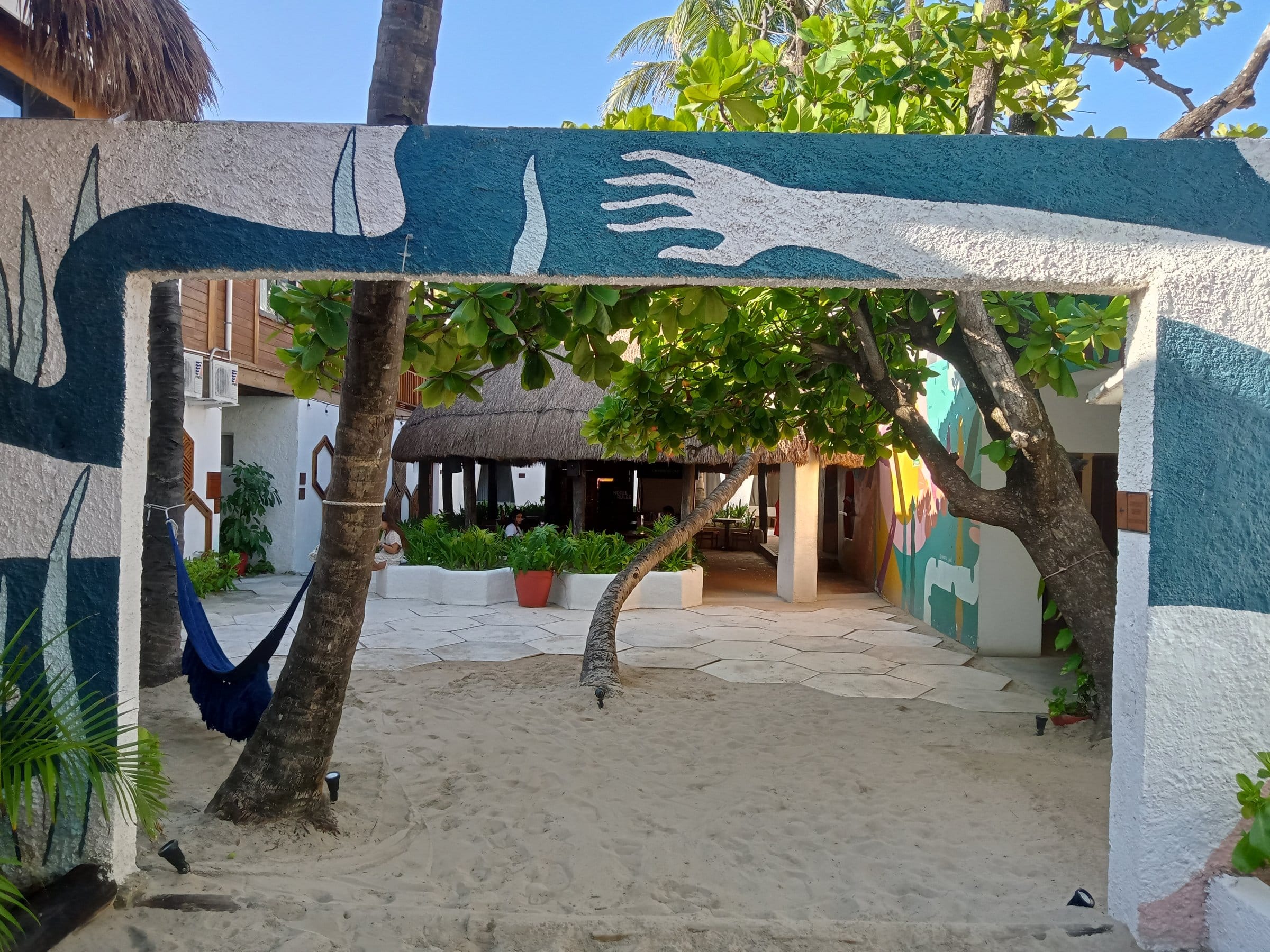 Courtyard entrance at Selina Isla Mujeres framed by vibrant murals and a hammock between trees with the beachfront palapa visible in the background