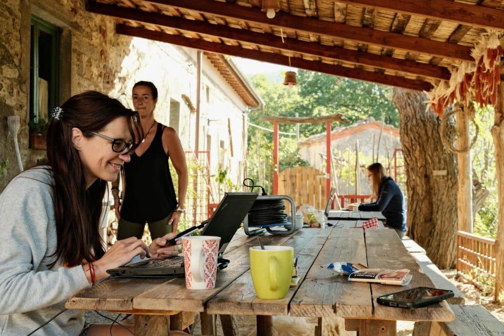 People working at laptops on a large wooden picnic table under a covered porch at a rustic stone building.