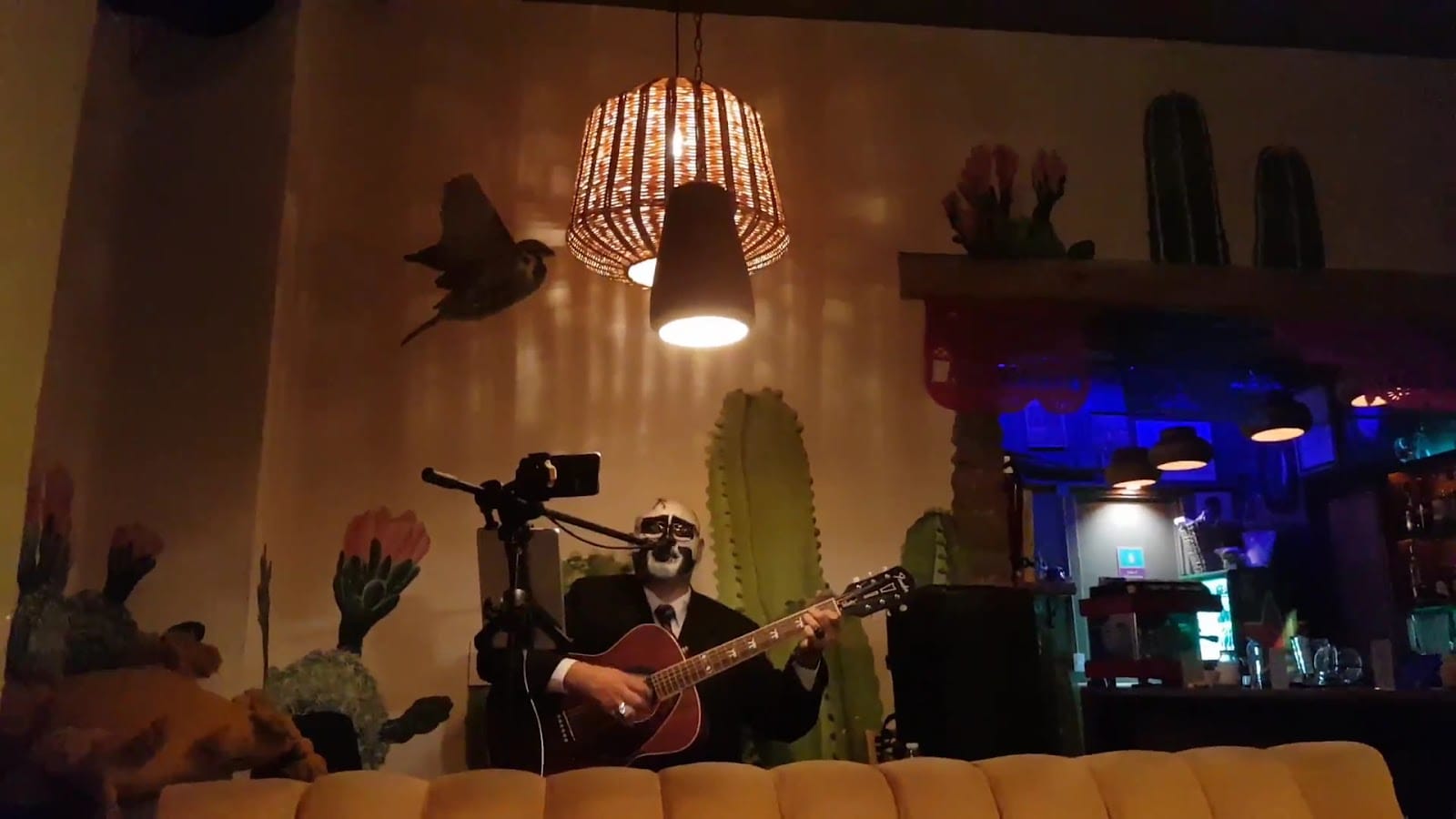 Live music event at Selina Oaxaca — a guitarist performing in front of a cactus mural in the bar area with guests watching from the yellow sofa