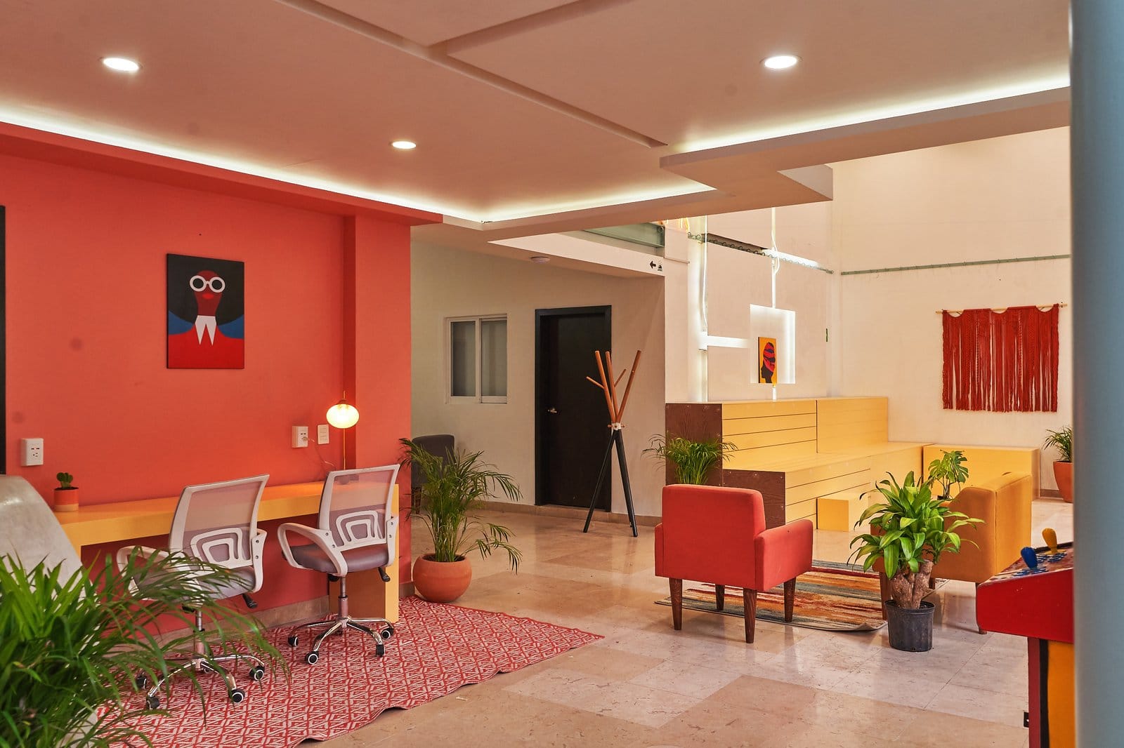 Coworking area at Nora Hotel Coliving Guadalajara — ergonomic mesh chairs and long desk against the red wall, warm lighting and plants throughout
