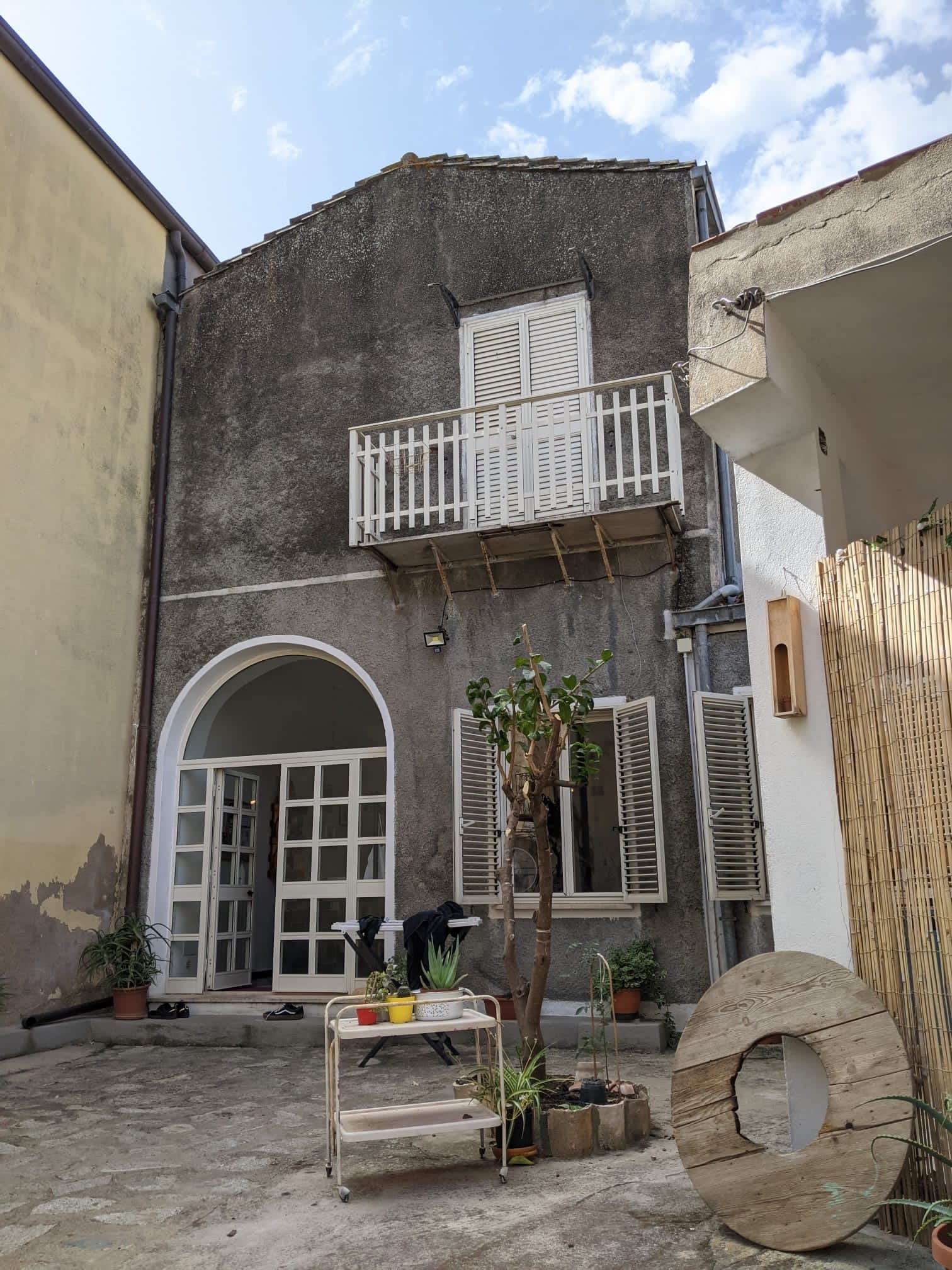 Rustic stone facade and sunlit courtyard with potted plants and small tree at Treballu Rural Coliving, an old village building in Laconi, Sardinia