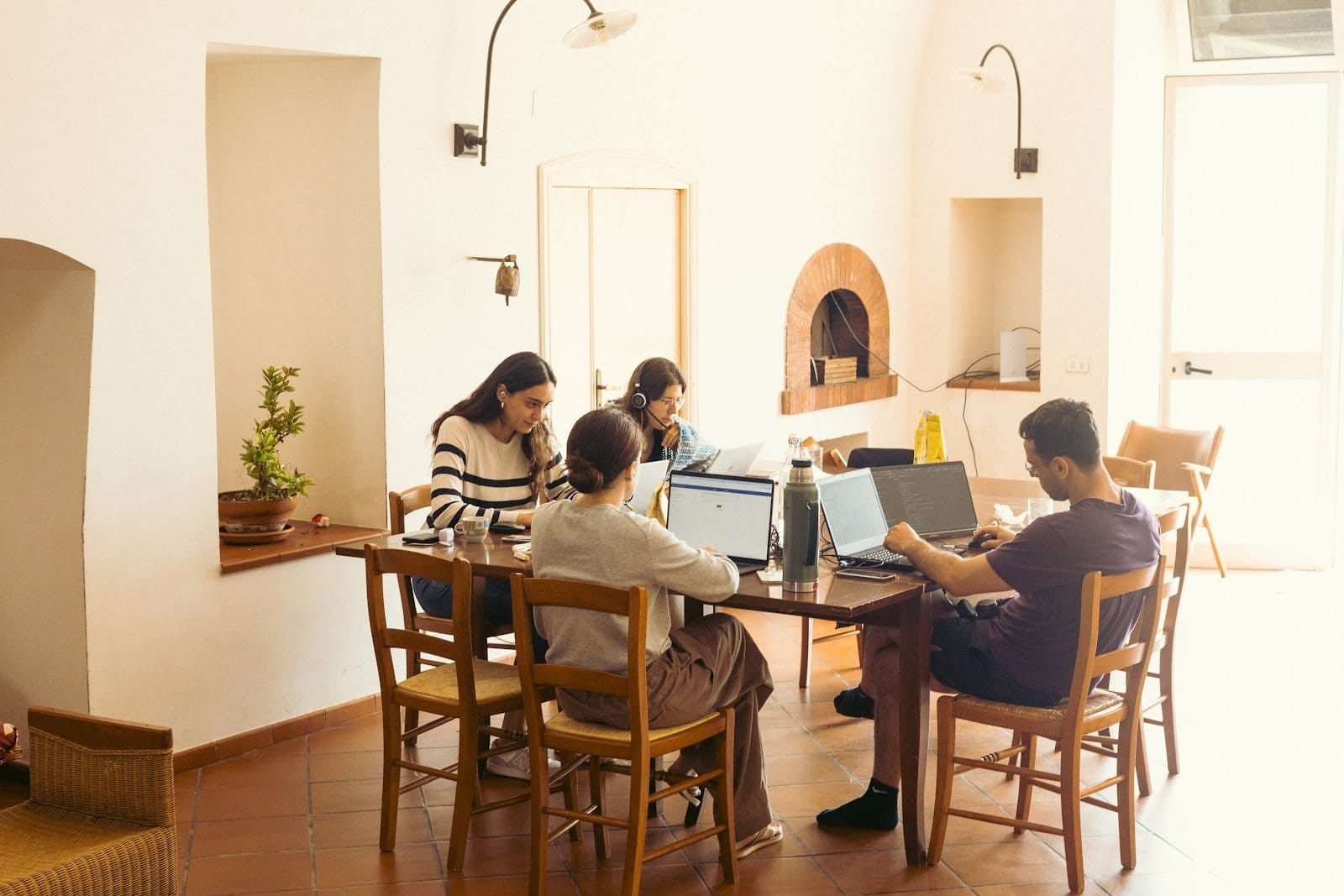 Warm indoor common dining area with four people sitting together at table using laptops and talking.