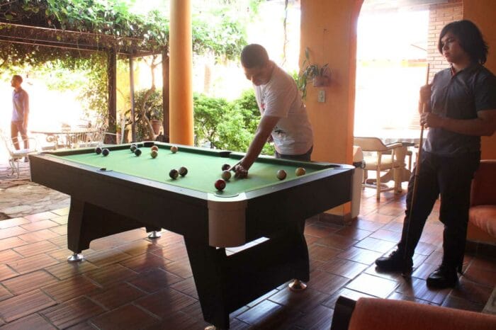 Several people playing billiards in a bright indoor common area with orange tile floor.