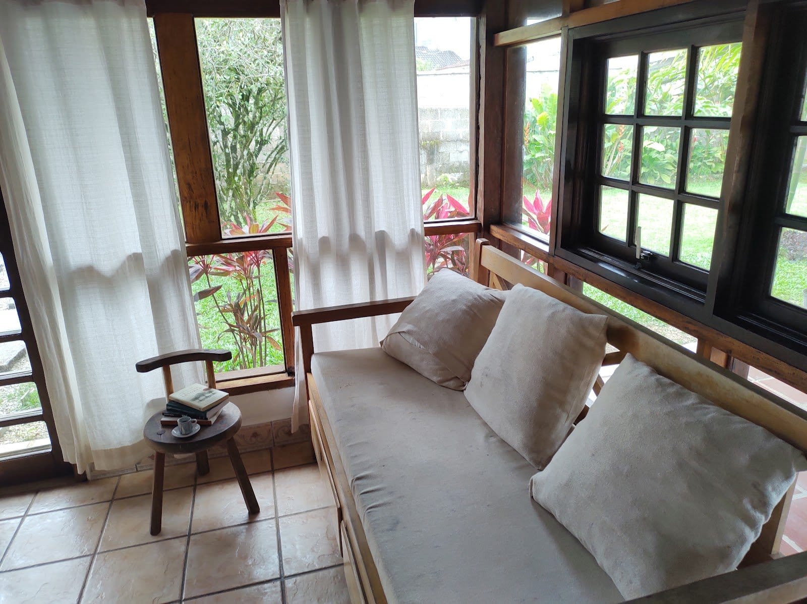 Bright reading nook with a cushioned wooden daybed and large windows looking onto the garden at Tô Aqui Casa de Vivências, Ubatuba