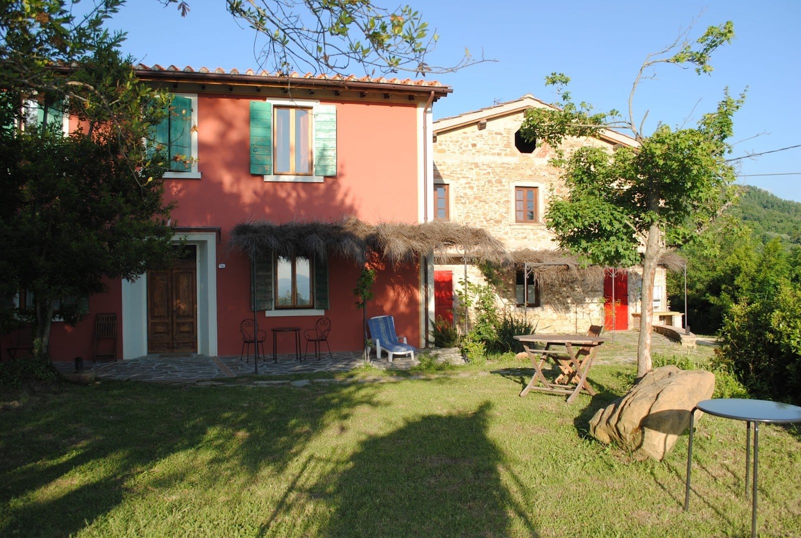 Sunlit stone house with red doors, surrounded by green lawn, trees, and garden furniture.