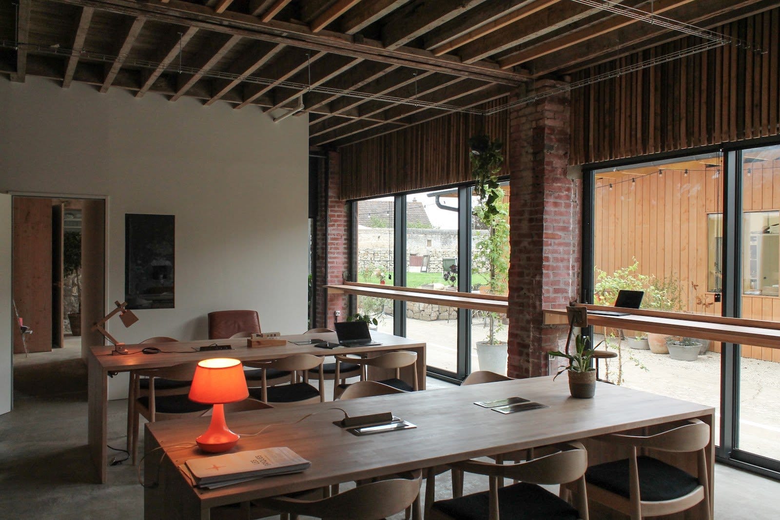 Indoor workspace with wooden desks, chairs, a lamp, and laptops by floor-to-ceiling windows opening to the garden.
