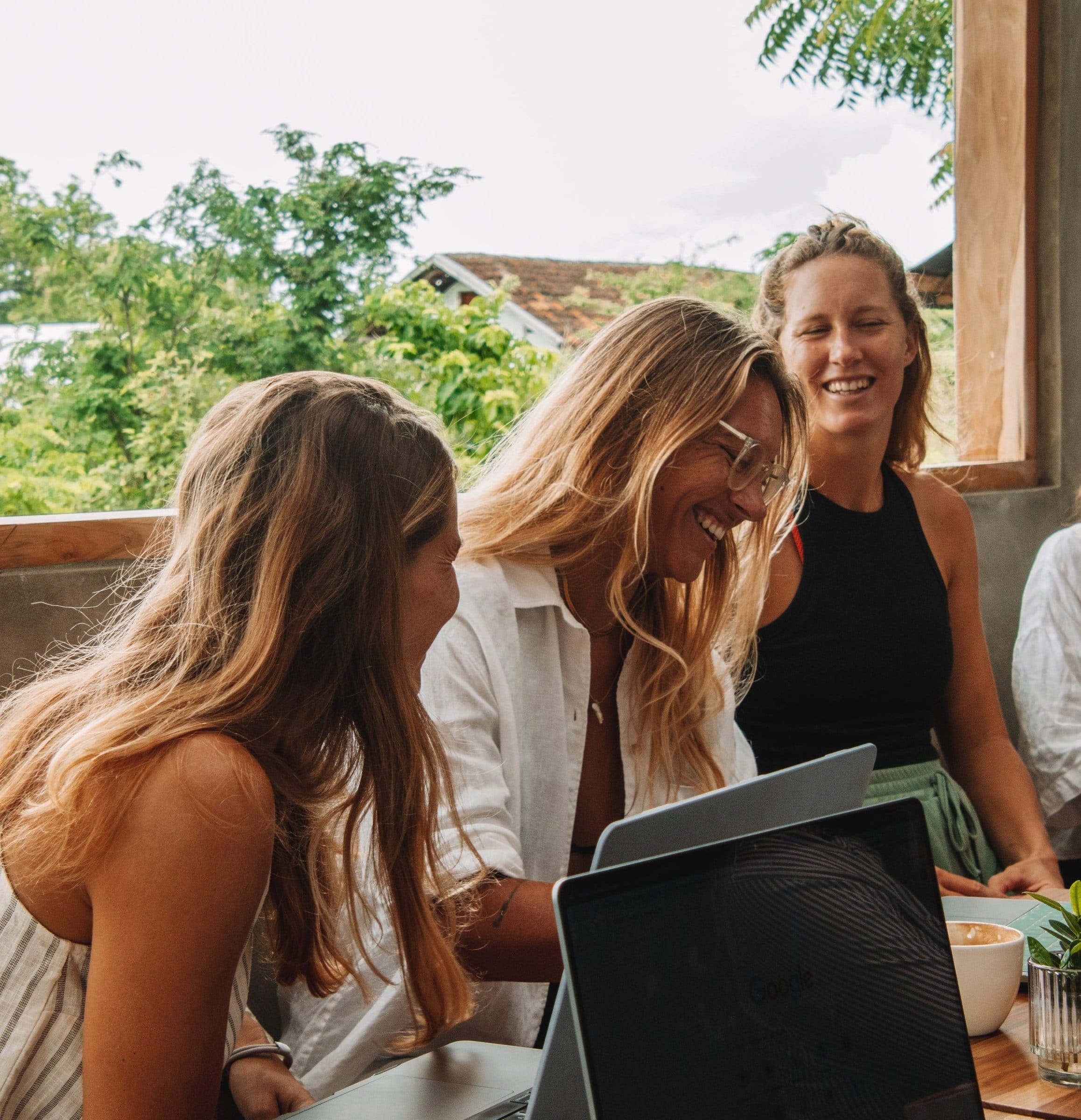 Group of three women laughing and socializing at a table with laptops and coffee in an indoor space with large windows.
