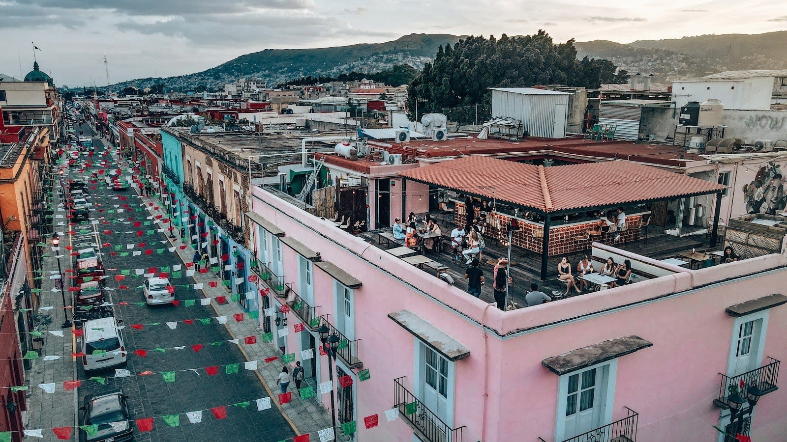 Aerial view of Selina Oaxaca at dusk showing the pink colonial building, rooftop terrace with guests, and the colourful Oaxacan street below strung with papel picado banners