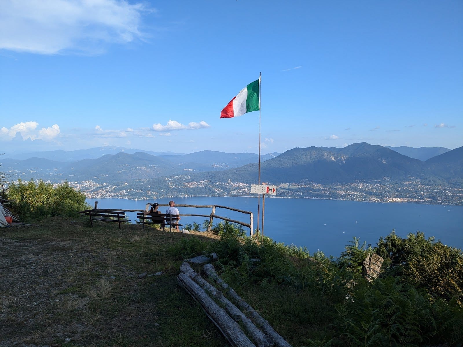 Two people sitting on benches in a grassy area with an Italian flag overhead and a panoramic lake and mountain view.