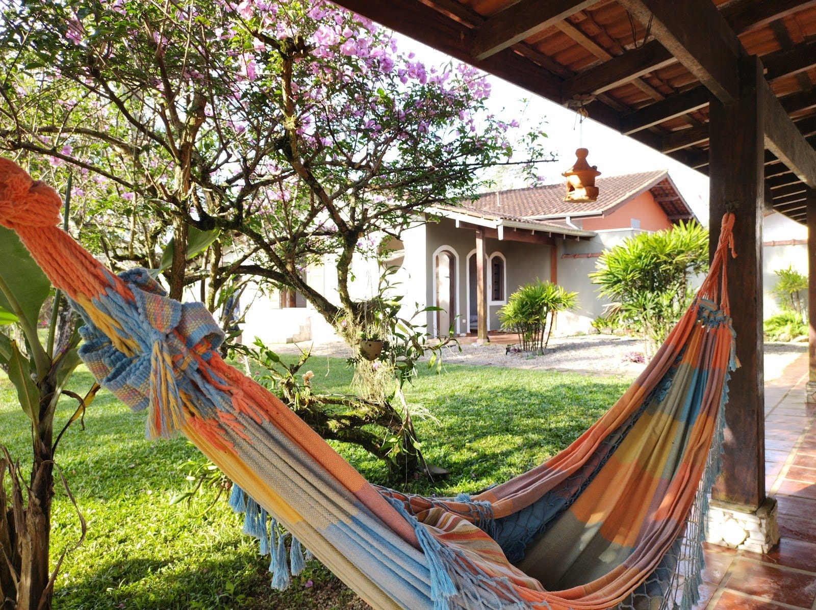 Colorful hammock under the wooden porch beside a flowering tree with the main house behind at Tô Aqui Casa de Vivências, Ubatuba