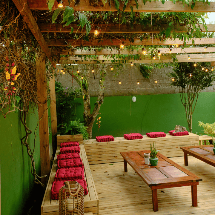 Covered wooden terrace with red-cushioned benches and string lights under a green trellis at Coliving Muní