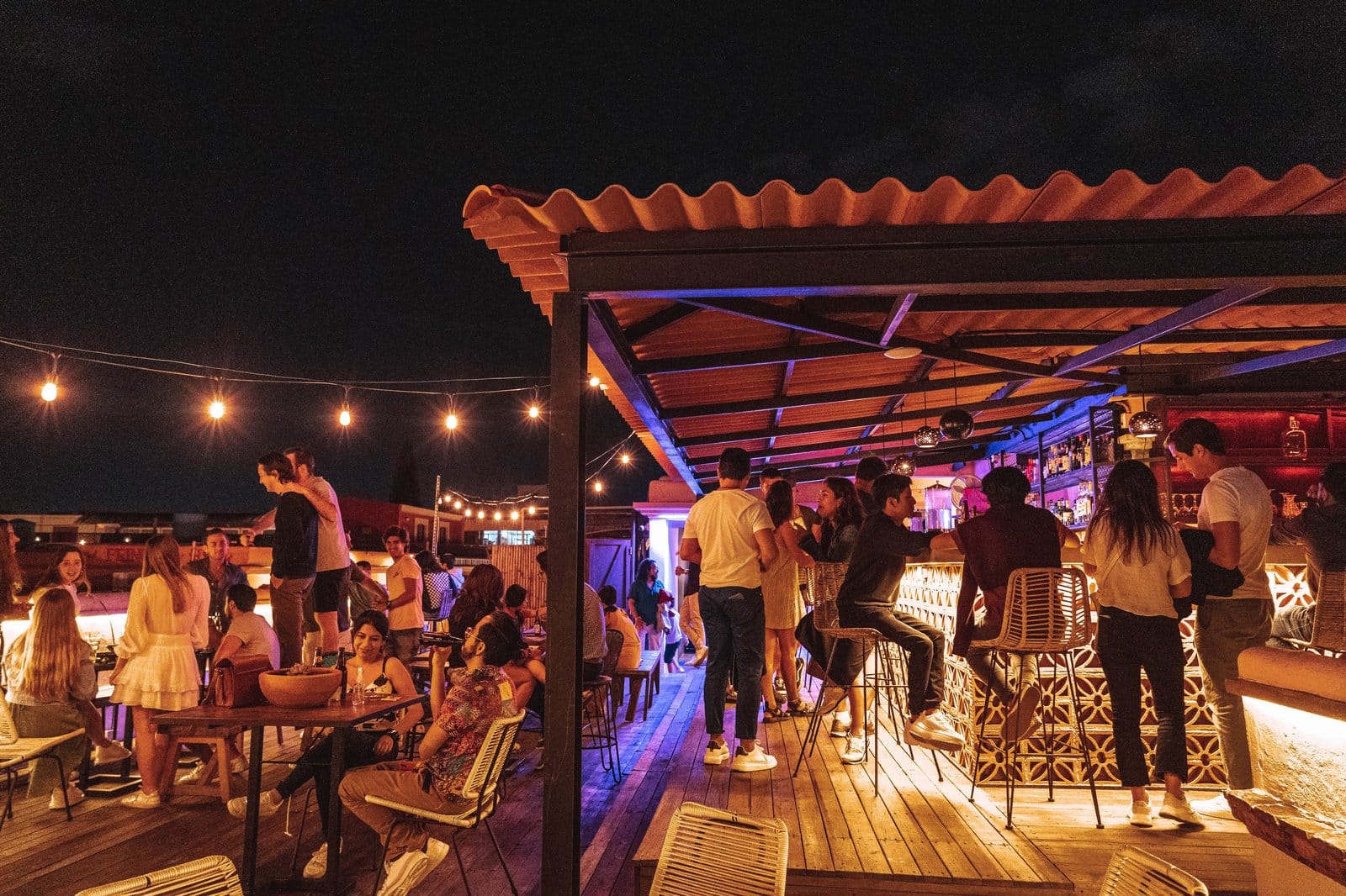 Lively rooftop bar at Selina Oaxaca at night with guests gathered under string lights around the open-air bar