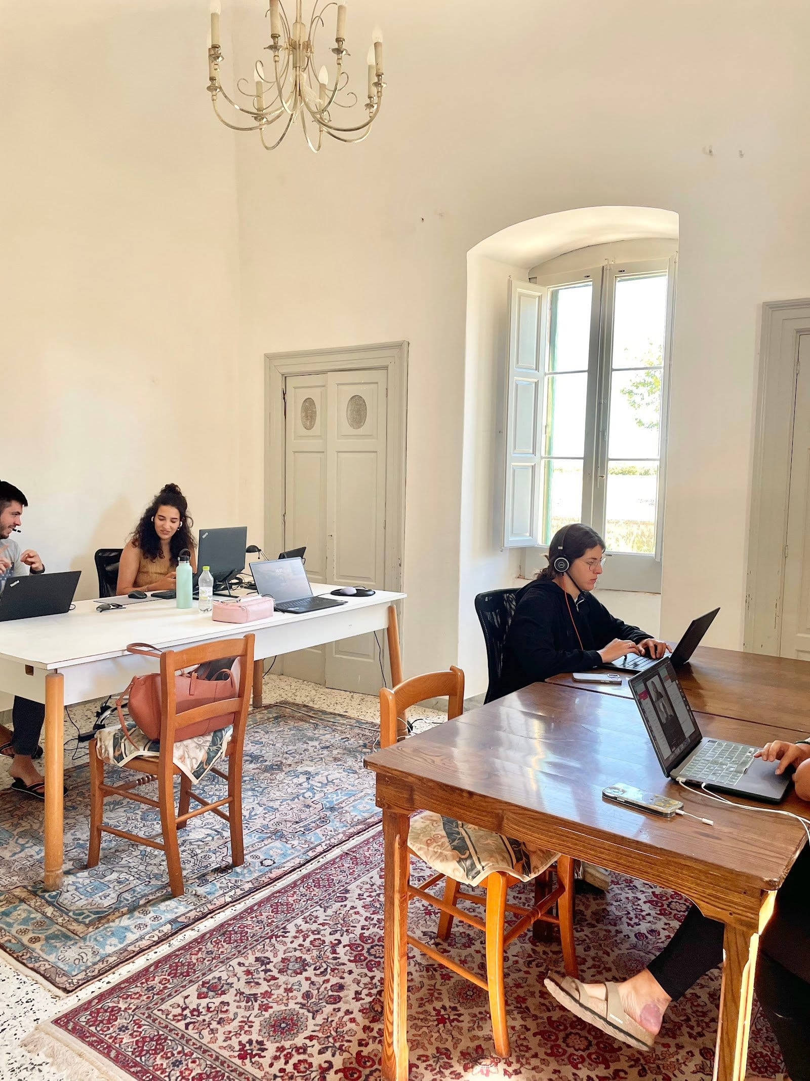 Bright indoor coworking space with multiple people seated at desks using laptops and conversing, white walls and open window.