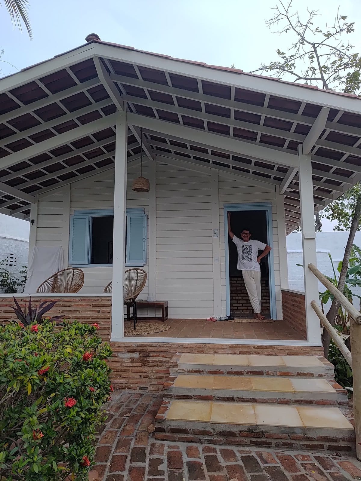 Guest on the porch of a white bungalow with rattan chairs at FlowHouse Jeri