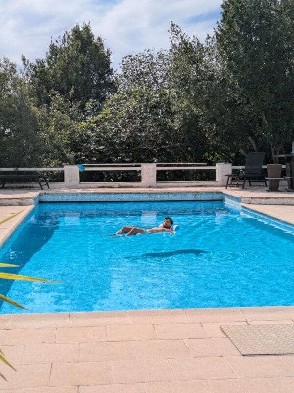 Person lying back floating on pool water with surrounding outdoor pool area visible on a sunny day.