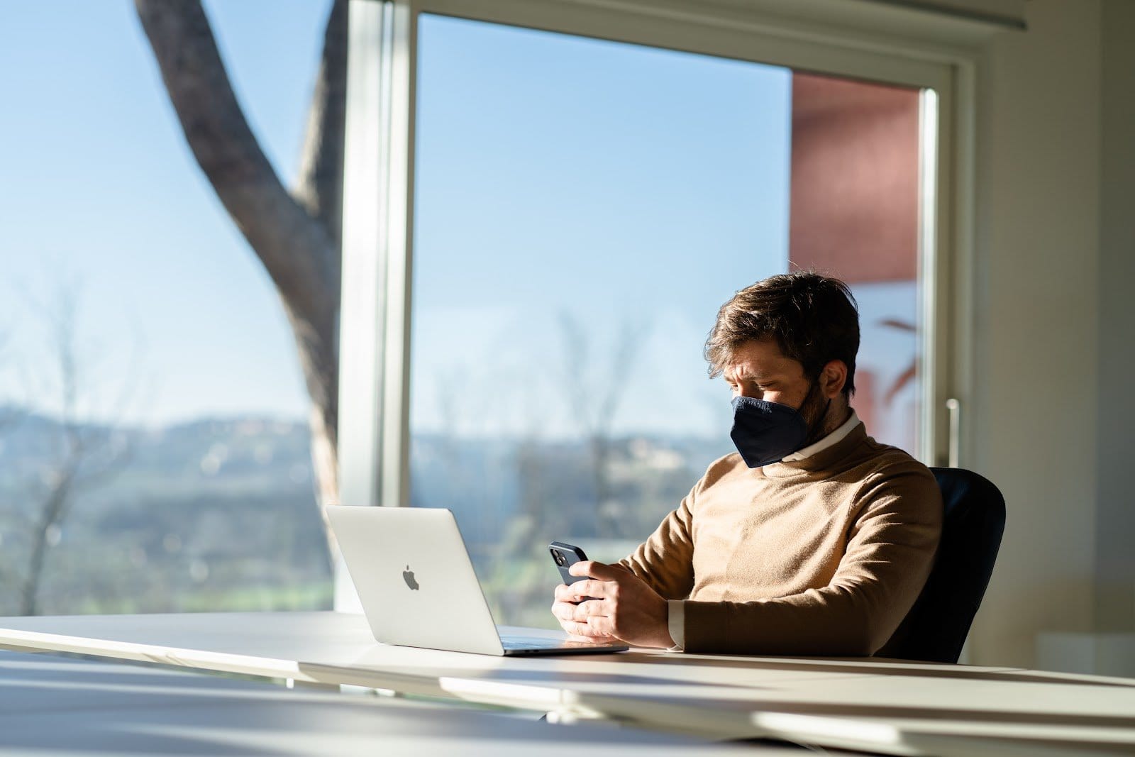 Bright indoor office space with person working at desk on laptop by large window with natural light.