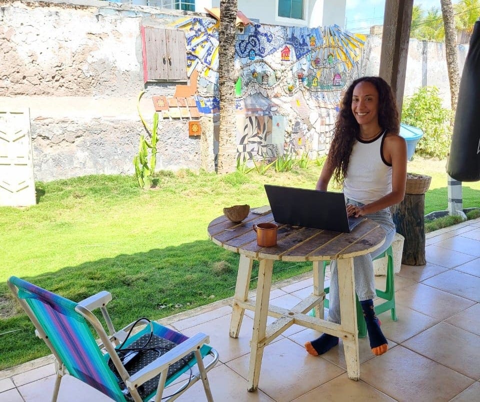 Resident smiling while working on a laptop at INFINIColiving covered patio in Camaçari, Brazil