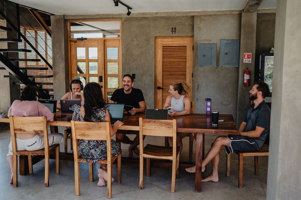 Several people working on laptops and chatting inside a well-lit communal coworking space with wooden furniture.