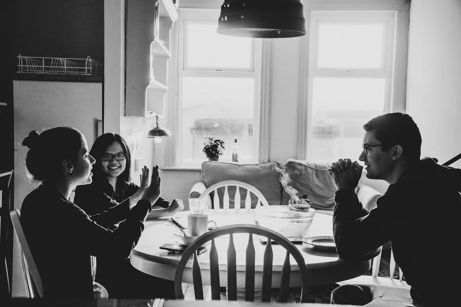 Group of three people sitting and smiling at a table in a cozy room with lamps and windows.