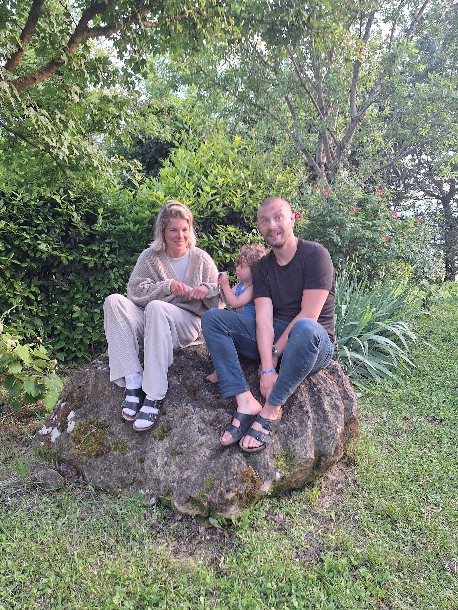 Three people sitting casually on a large rock outdoors, smiling and interacting socially.