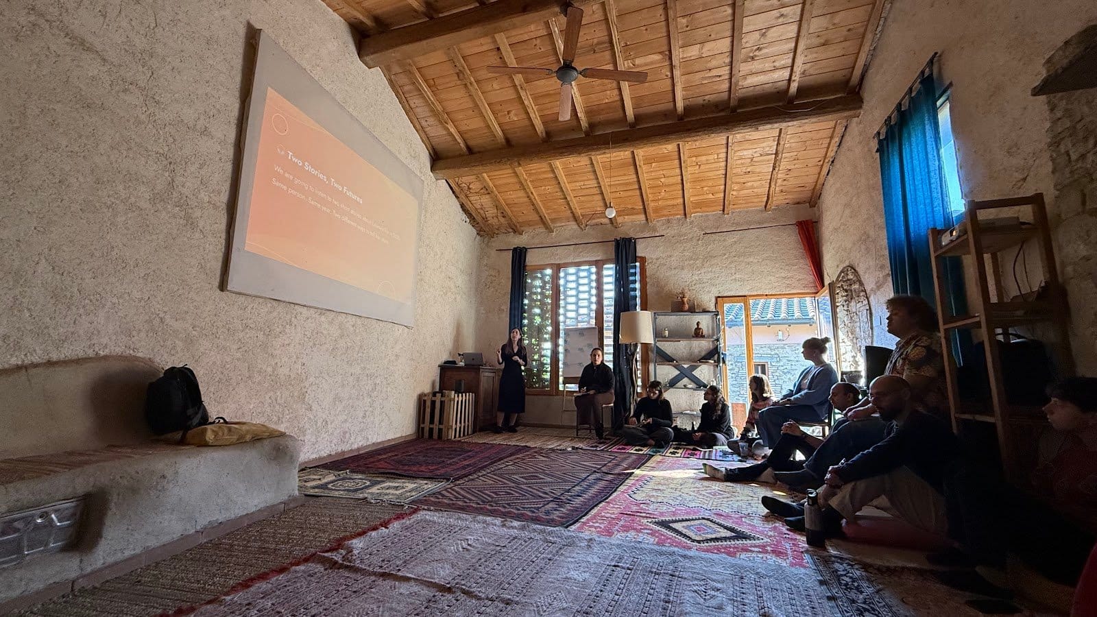 A wide interior room with rustic walls and a wooden ceiling. Several people are seated on floor cushions and chairs in front of a projected presentation, indicating a group activity or meeting.