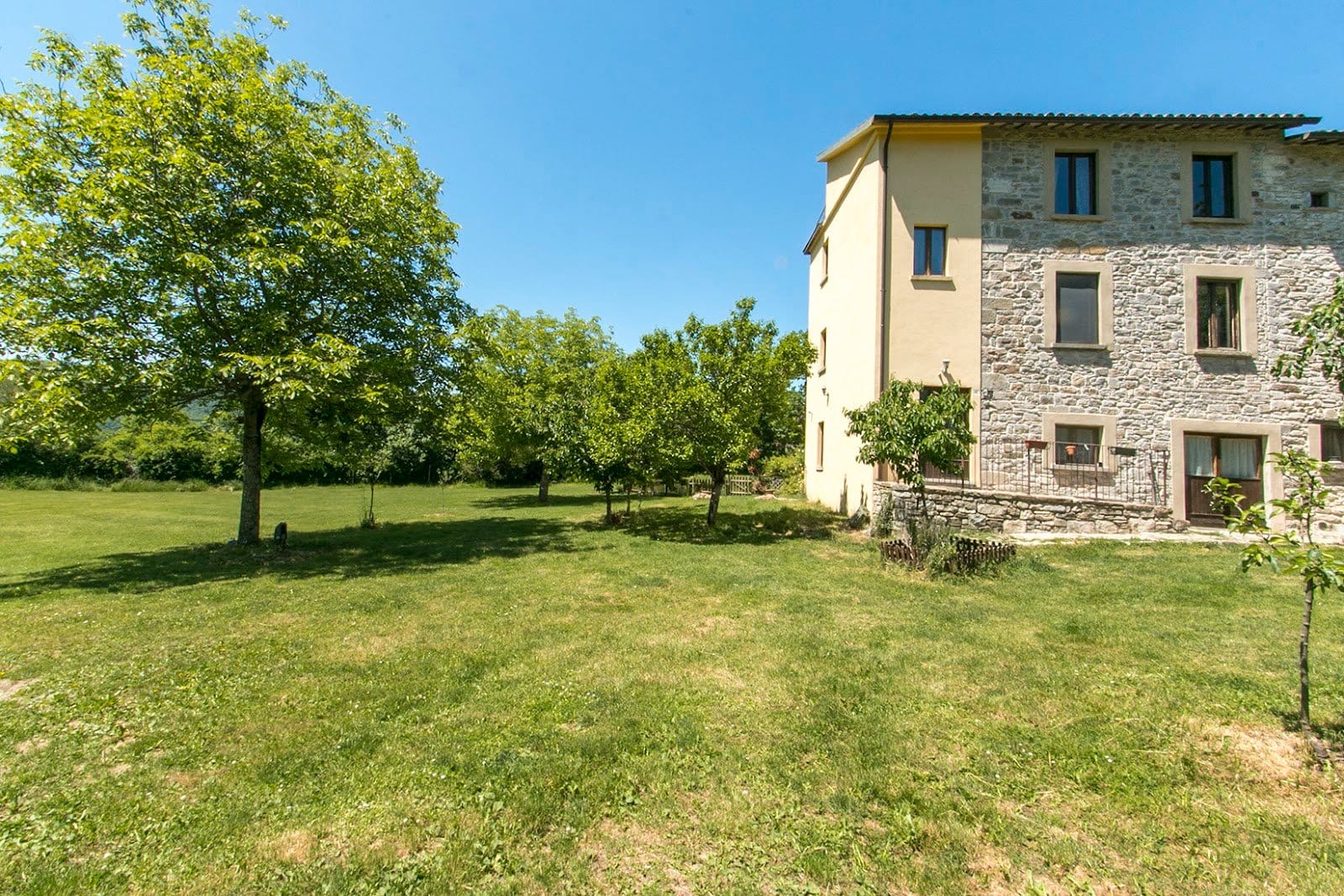 Outdoor photo showing a side of a stone and plaster building next to a grassy garden with mature trees under a clear blue sky.
