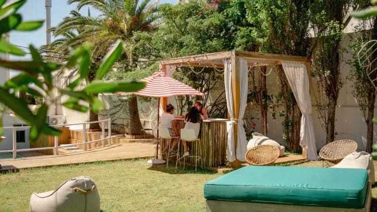 Outdoor garden area with greenery, canopy, wicker chairs, beanbags, and people sitting and chatting at a high table.