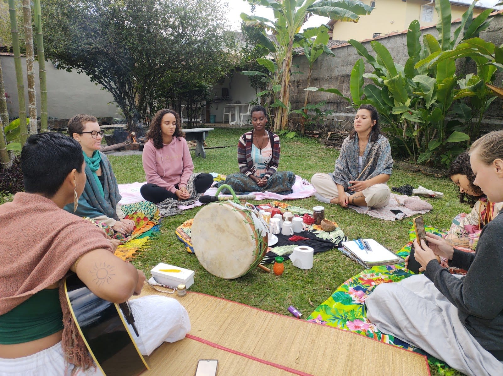 Guests gathered in a circle on the garden lawn with a drum and guitar during a community gathering at Tô Aqui Casa de Vivências, Ubatuba