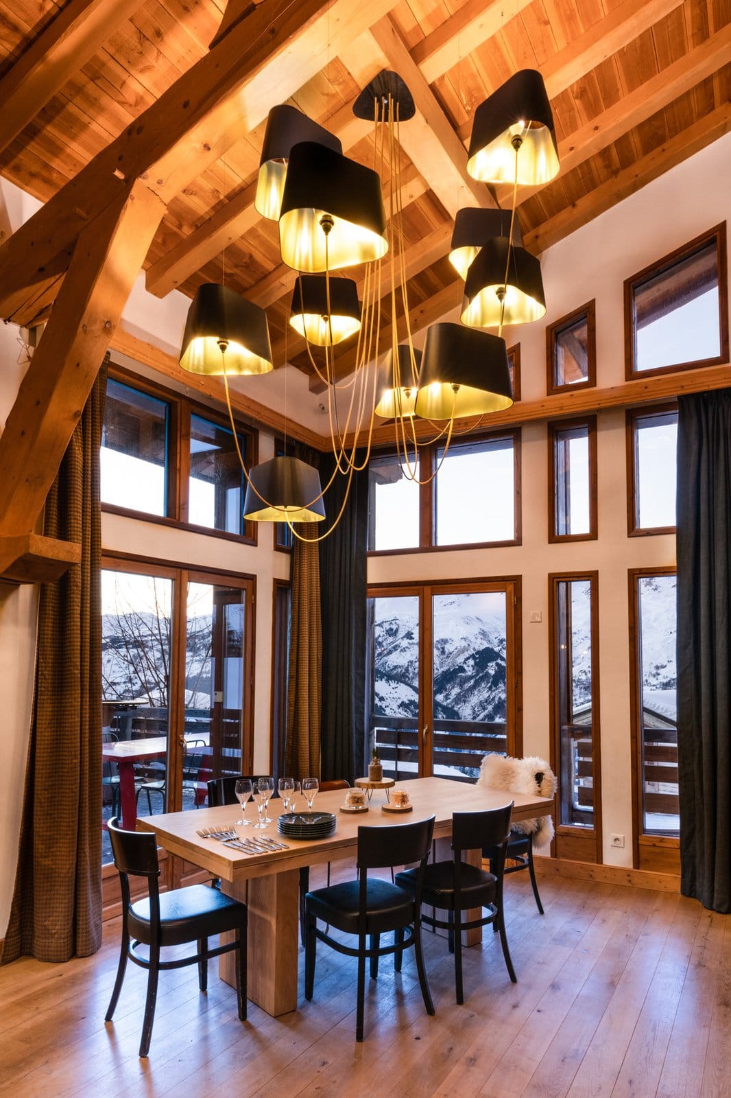 Elegant indoor dining room with wooden table and black chairs under multiple modern black ceiling lamps, large windows revealing snowy outdoor scenery.