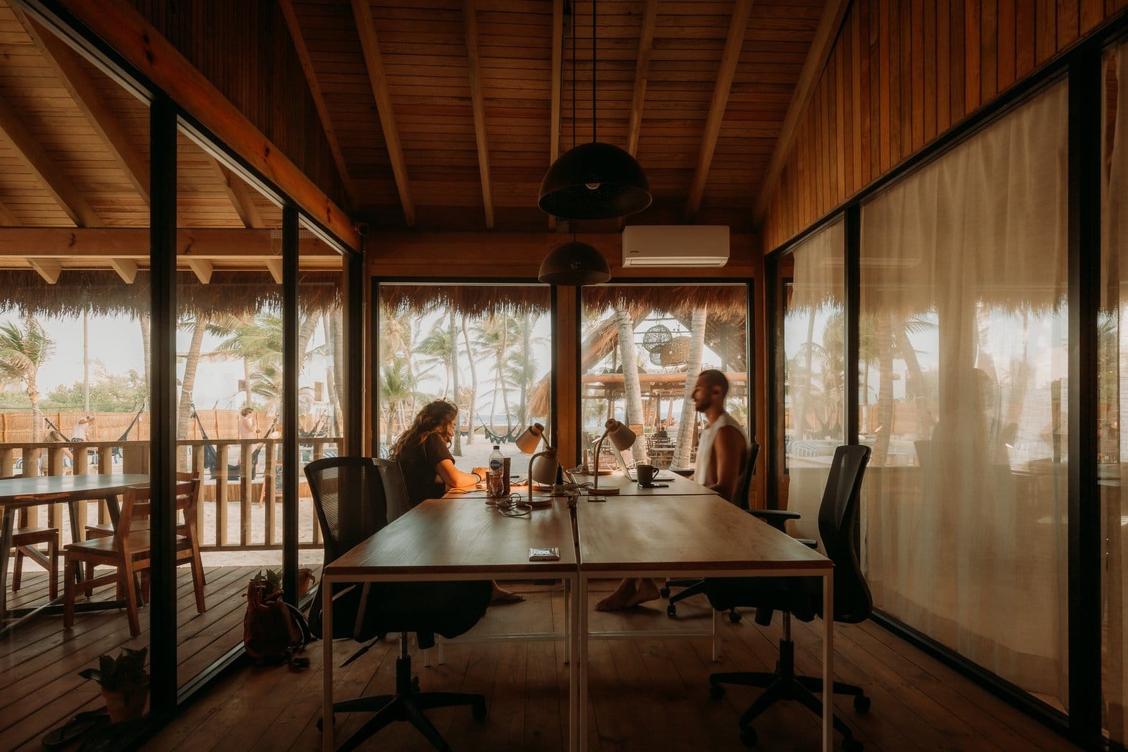 Two residents working in the glass-walled meeting room at Selina Isla Mujeres with palm trees and beach visible outside