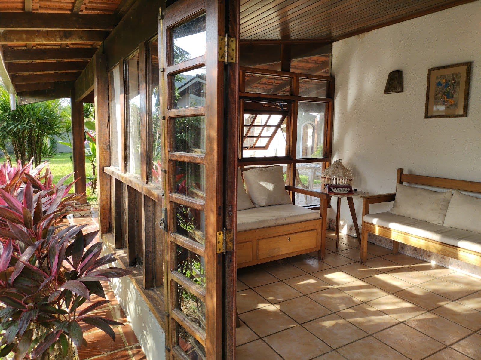 Sunny enclosed wooden veranda lounge with daybeds and glass doors opening to the garden at Tô Aqui Casa de Vivências, Ubatuba