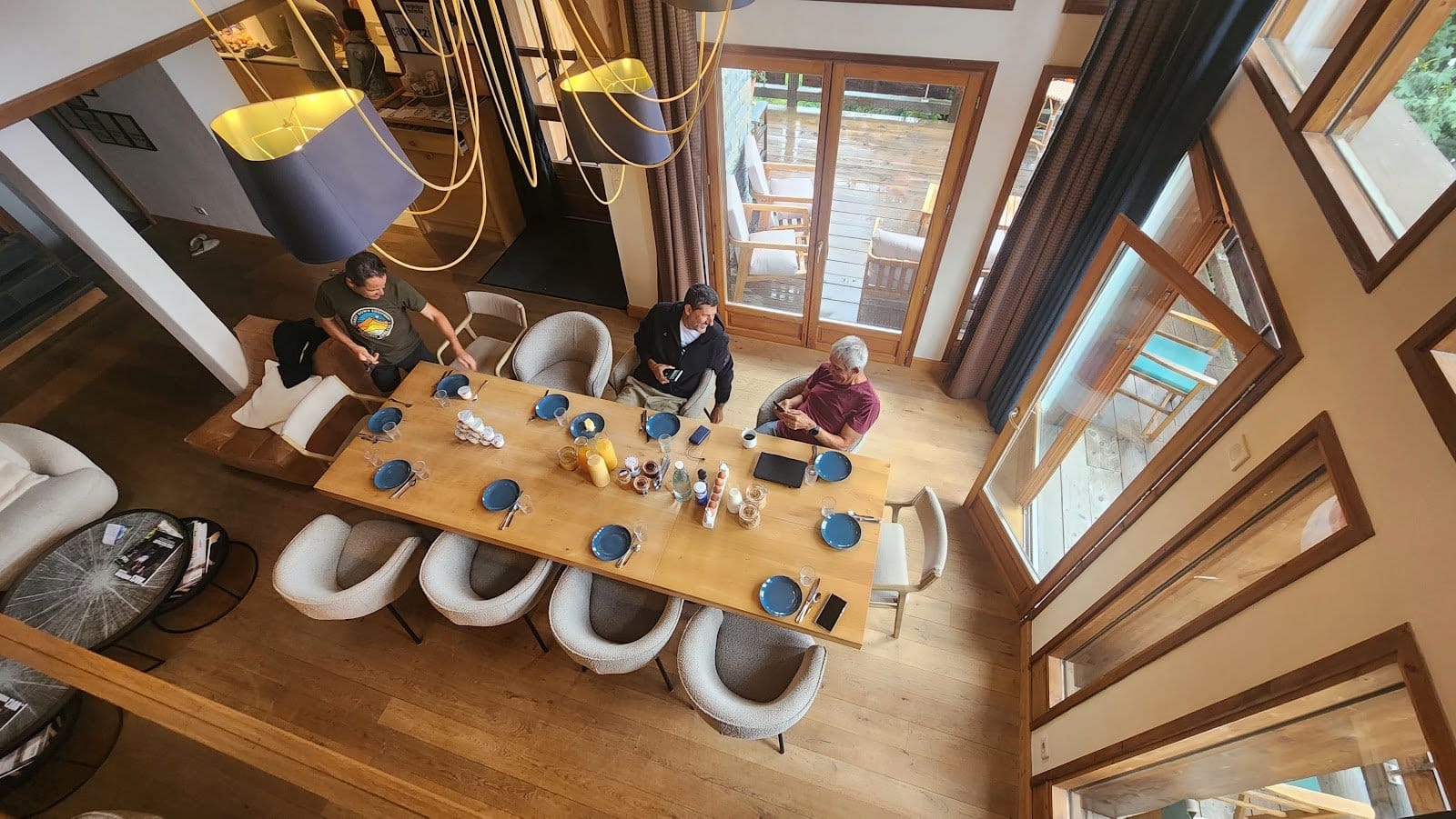 Overhead view of people gathered around a large wooden dining table enjoying a meal in a bright, airy room with lots of windows.