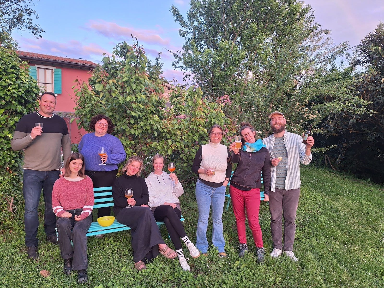 Eight people standing and sitting outdoors in a garden with drinks, smiling and enjoying social gathering in evening light.